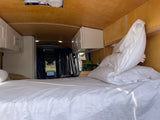 Cozy RV rental interior showing a comfortable bed with white linens, overhead cabinets, and front views.