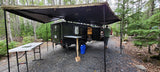 An off-road rv rental camper with an extended awning in a forest. A man stands near its open rear hatch, with camping gear visible.