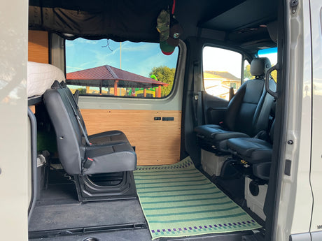 Interior of a camper van RV rental, showing black seats, wooden wall panels, a green rug, and an outside park view.