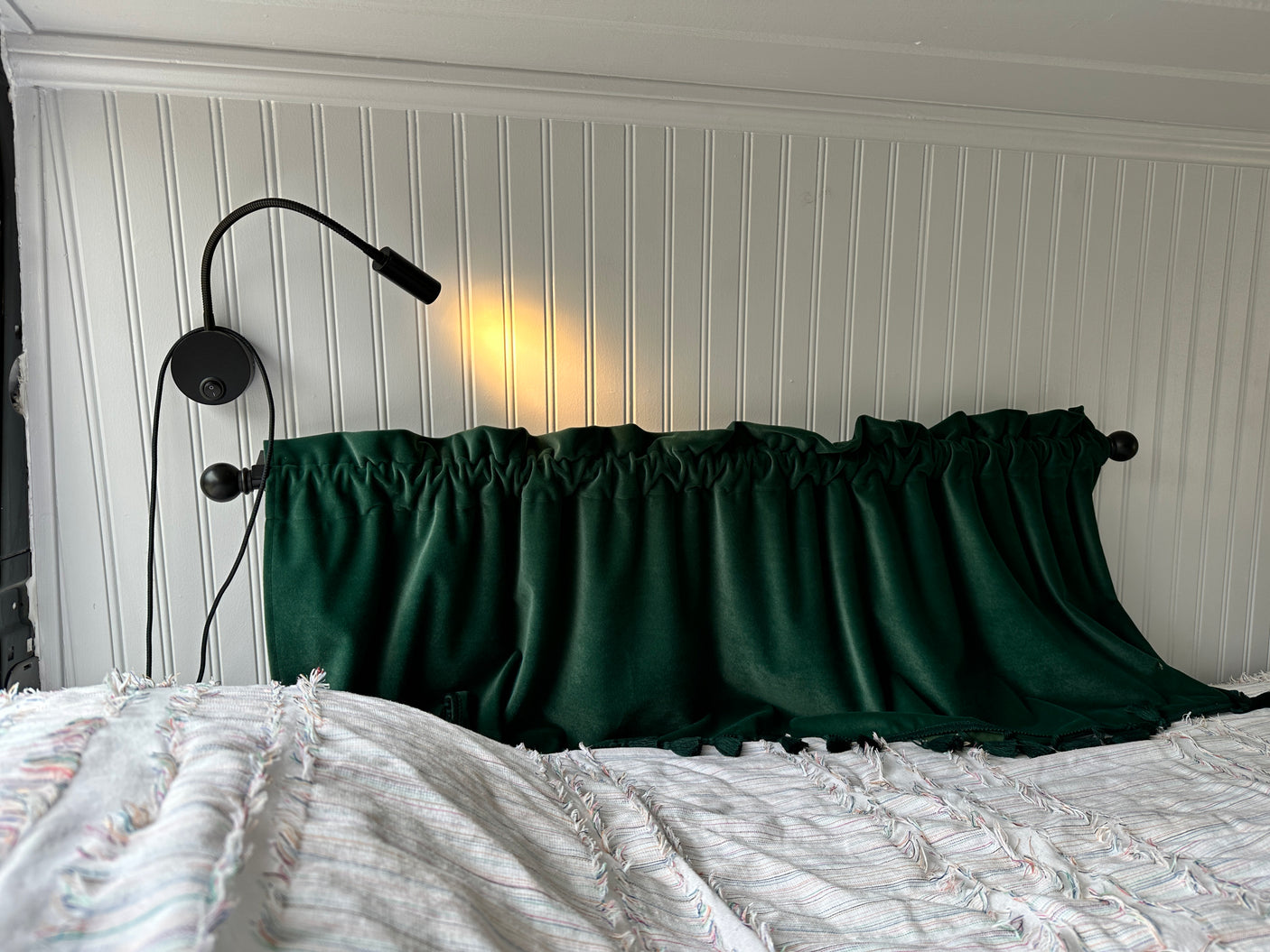 Cozy RV rental bedroom: white beadboard wall, reading lamp, and dark green velvet valance over a comfortable bed.