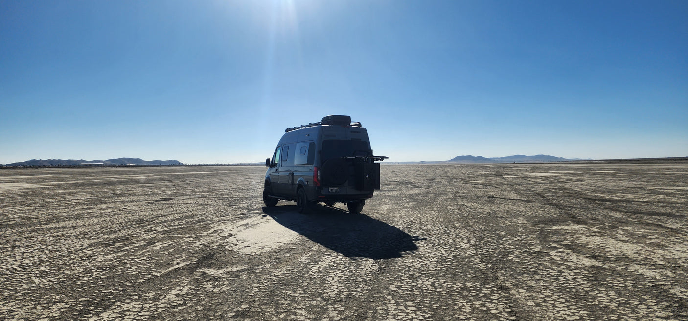 Grey camper van (RV rental) on a vast cracked desert floor under a clear blue sky, with distant mountains.