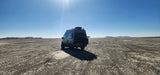 Grey camper van (RV rental) on a vast cracked desert floor under a clear blue sky, with distant mountains.
