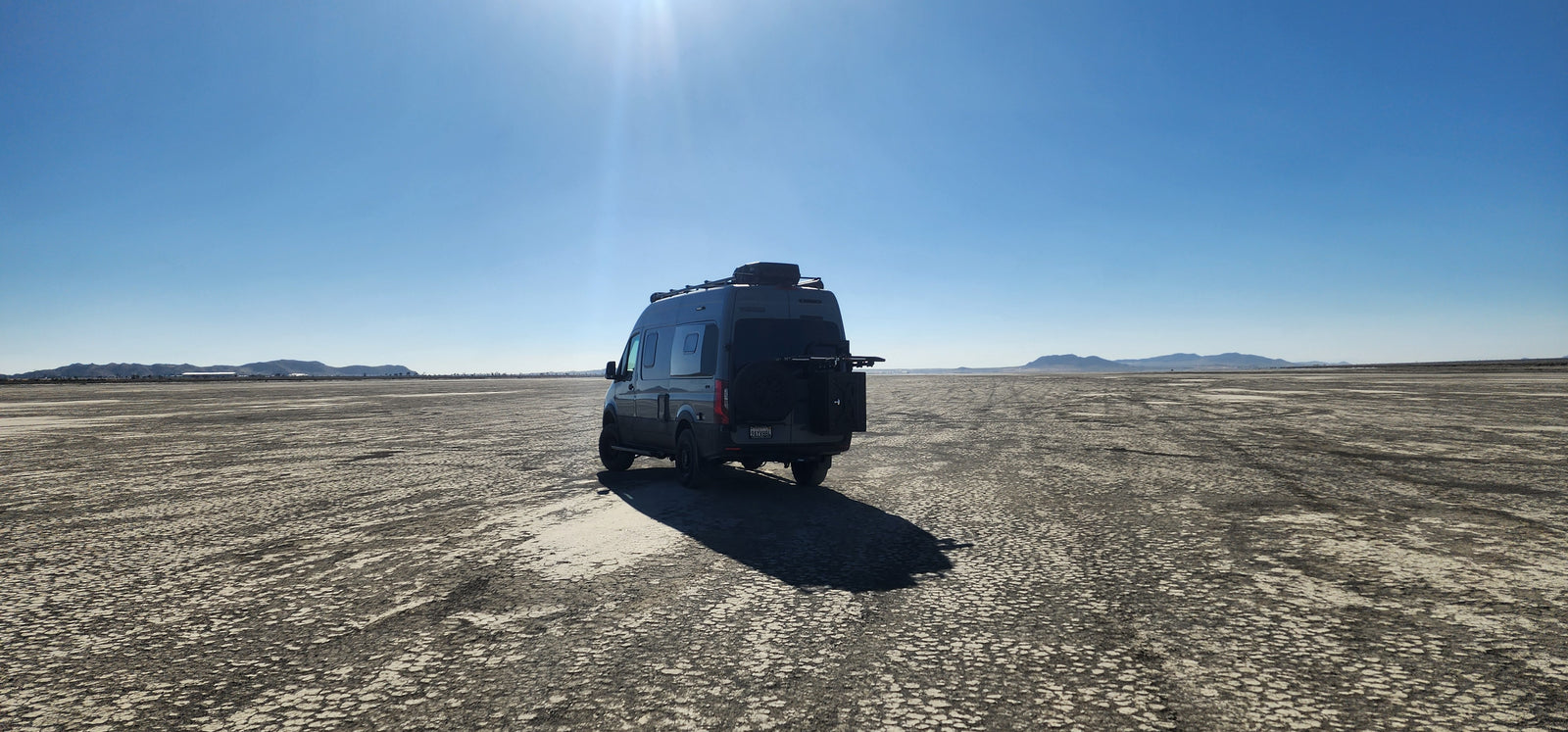 Grey camper van (RV rental) on a vast cracked desert floor under a clear blue sky, with distant mountains.