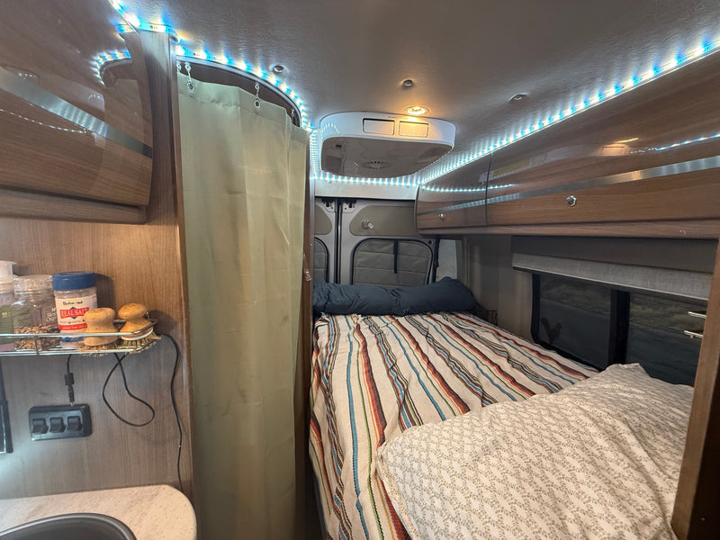 Cozy RV rental interior with bed, overhead cabinets, LED lighting, and a compact kitchen area. — photo 10