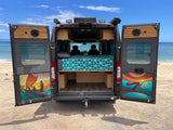 This RV rental is parked on a beach with its back doors open, revealing a cozy bed and ocean views. Doors feature vibrant art.