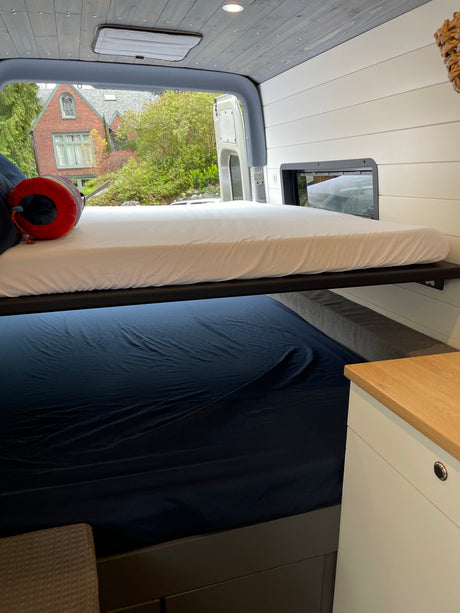 Modern RV rental interior with bunk beds, white and navy bedding, and an outside view from the open rear doors.
