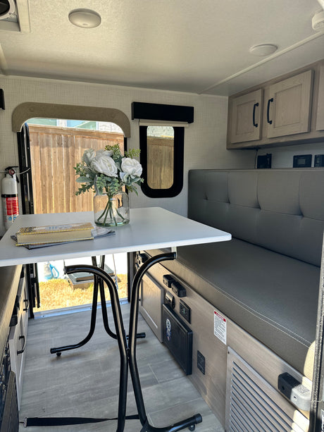 Modern rv rental dinette area with white table, gray bench, and overhead cabinets. Window shows outdoor fence.