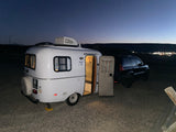 A white Skyler teardrop RV rental with its door open, hitched to a dark SUV, parked on gravel at dusk with distant city lights.