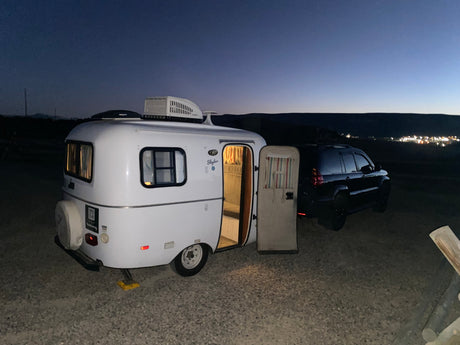 A white Skyler teardrop RV rental with its door open, hitched to a dark SUV, parked on gravel at dusk with distant city lights.