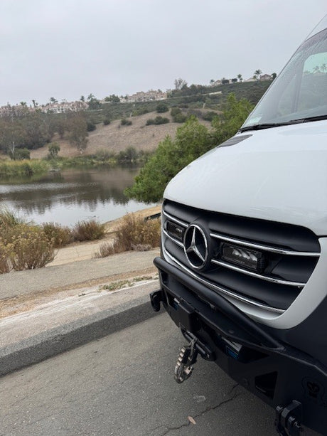 White Mercedes-Benz Sprinter van, ideal for RV rental adventures, parked by a scenic lake with hills and homes.