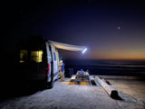 Night beach camping with an RV rental. Person near van's lit awning, grill on table by the ocean under stars.