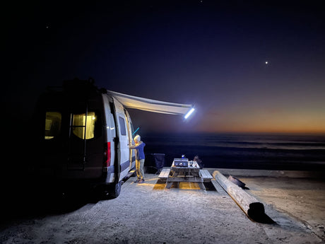 Night beach camping with an RV rental. Person near van's lit awning, grill on table by the ocean under stars.