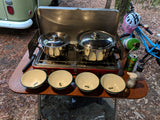 An RV rental campsite view: two pots on a camp stove, with four bowls, spices, and a beer on a wooden table. A bike and vintage RV are visible.