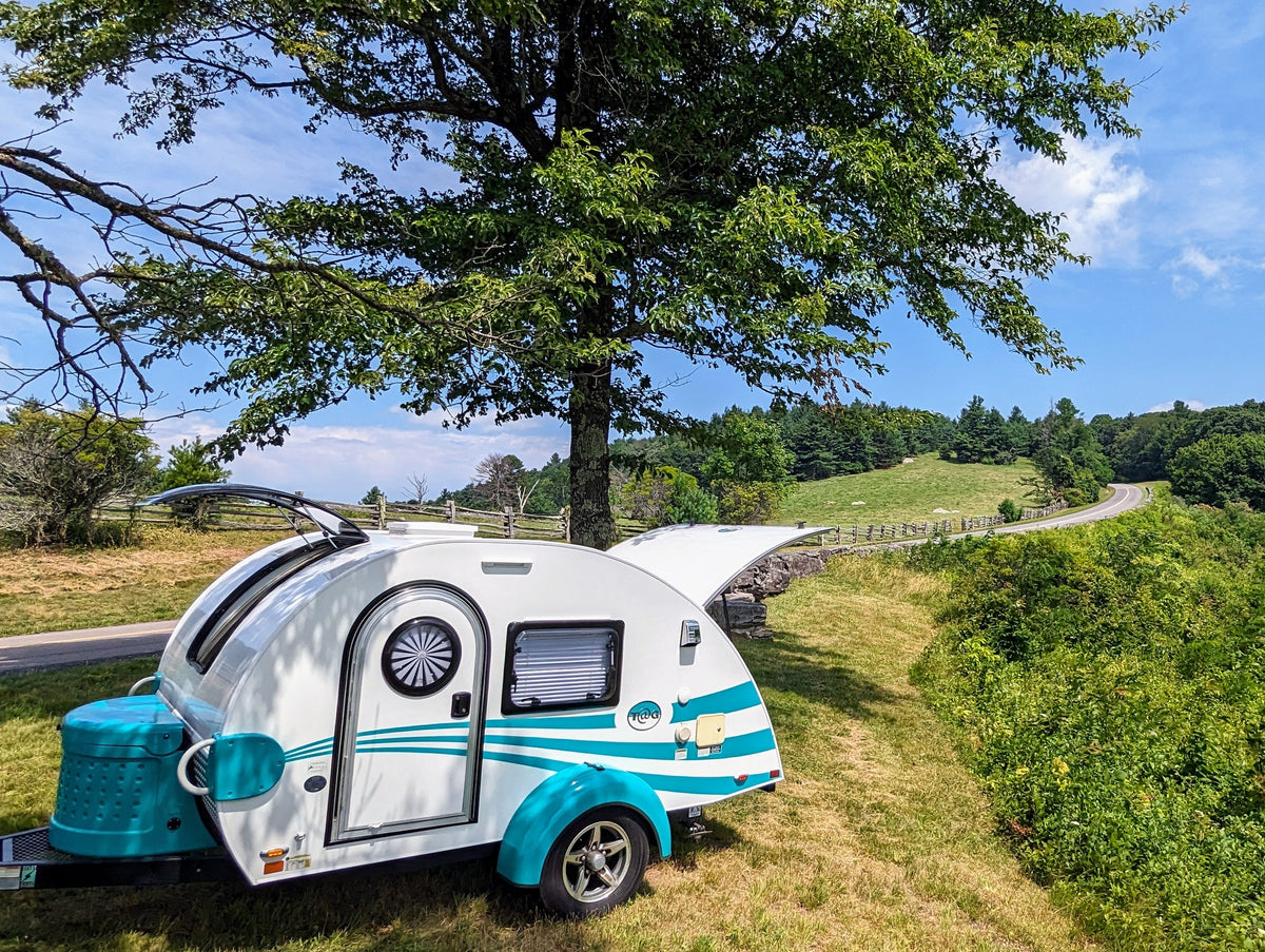Scenic RV rental adventure! A white & teal teardrop camper under a tree, with a winding road & green hills in the background. — photo 17