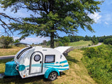 Scenic RV rental adventure! A white & teal teardrop camper under a tree, with a winding road & green hills in the background.
