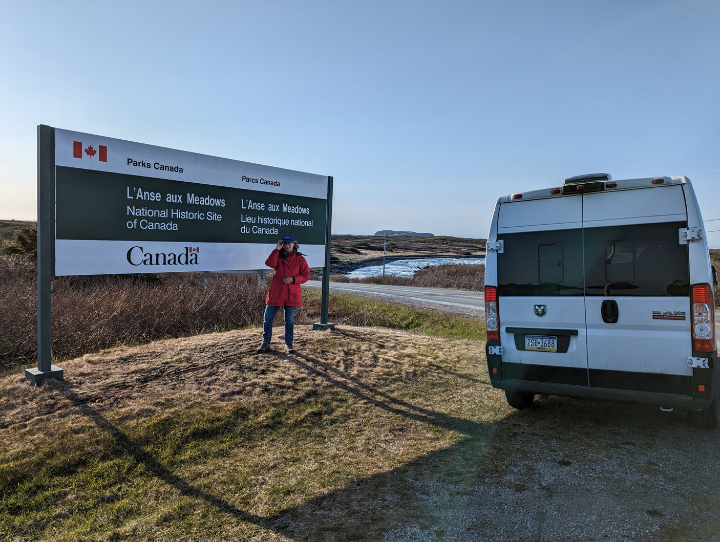 A person in a red jacket stands by the L'Anse aux Meadows National Historic Site sign. Their white RV rental is parked on the right.