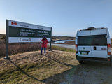 A person in a red jacket stands by the L'Anse aux Meadows National Historic Site sign. Their white RV rental is parked on the right.