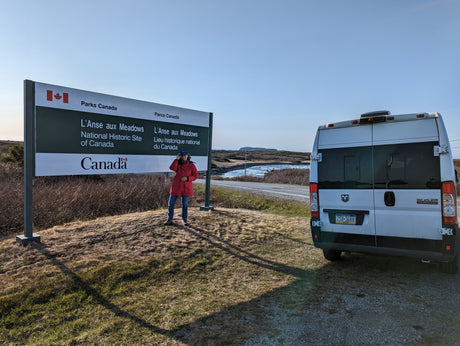 A person in a red jacket stands by the L'Anse aux Meadows National Historic Site sign. Their white RV rental is parked on the right.