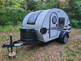 This compact RV rental is a white teardrop trailer parked in a serene wooded campsite.