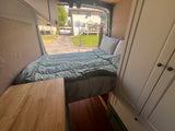 RV rental van interior: comfortable bed, pillows, wooden cabinetry, and open rear doors revealing a residential driveway view.