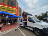 White RV rental van with open interior & rooftop tent parked at Boulder Theater for Teton Gravity Research event. People gather outside.