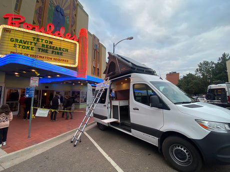 White RV rental van with open interior & rooftop tent parked at Boulder Theater for Teton Gravity Research event. People gather outside.