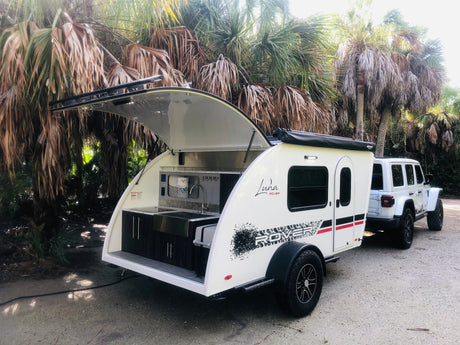 This RV rental features a white teardrop trailer with an open outdoor kitchen, hitched to a Jeep, parked by palm trees.