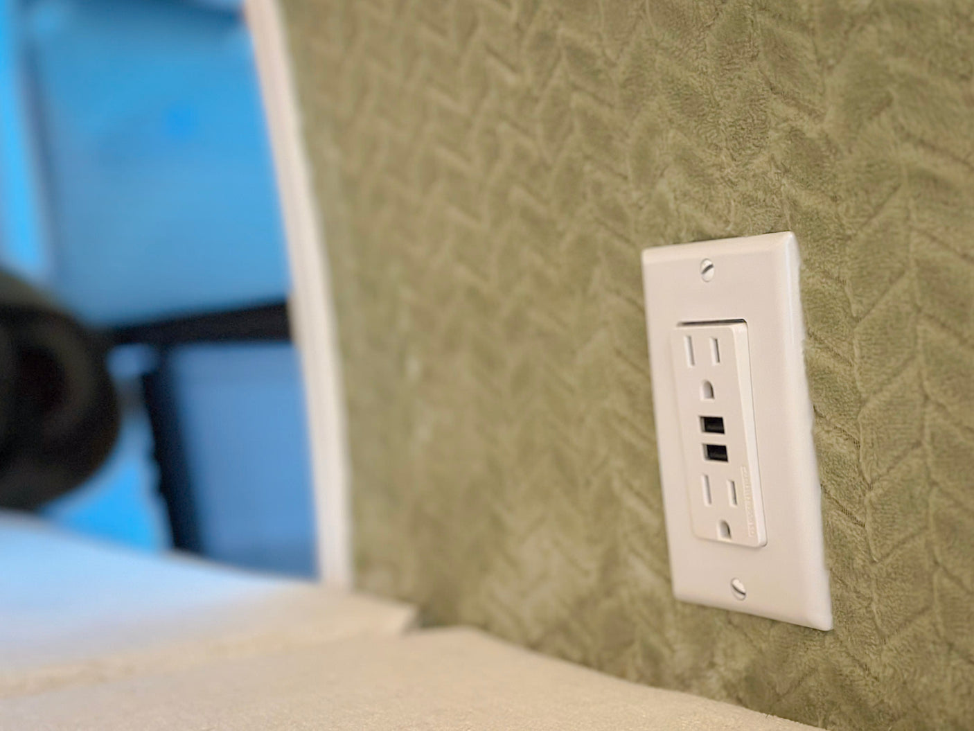 RV rental interior: Close-up of a white outlet offering AC and USB power on a soft, patterned green wall.
