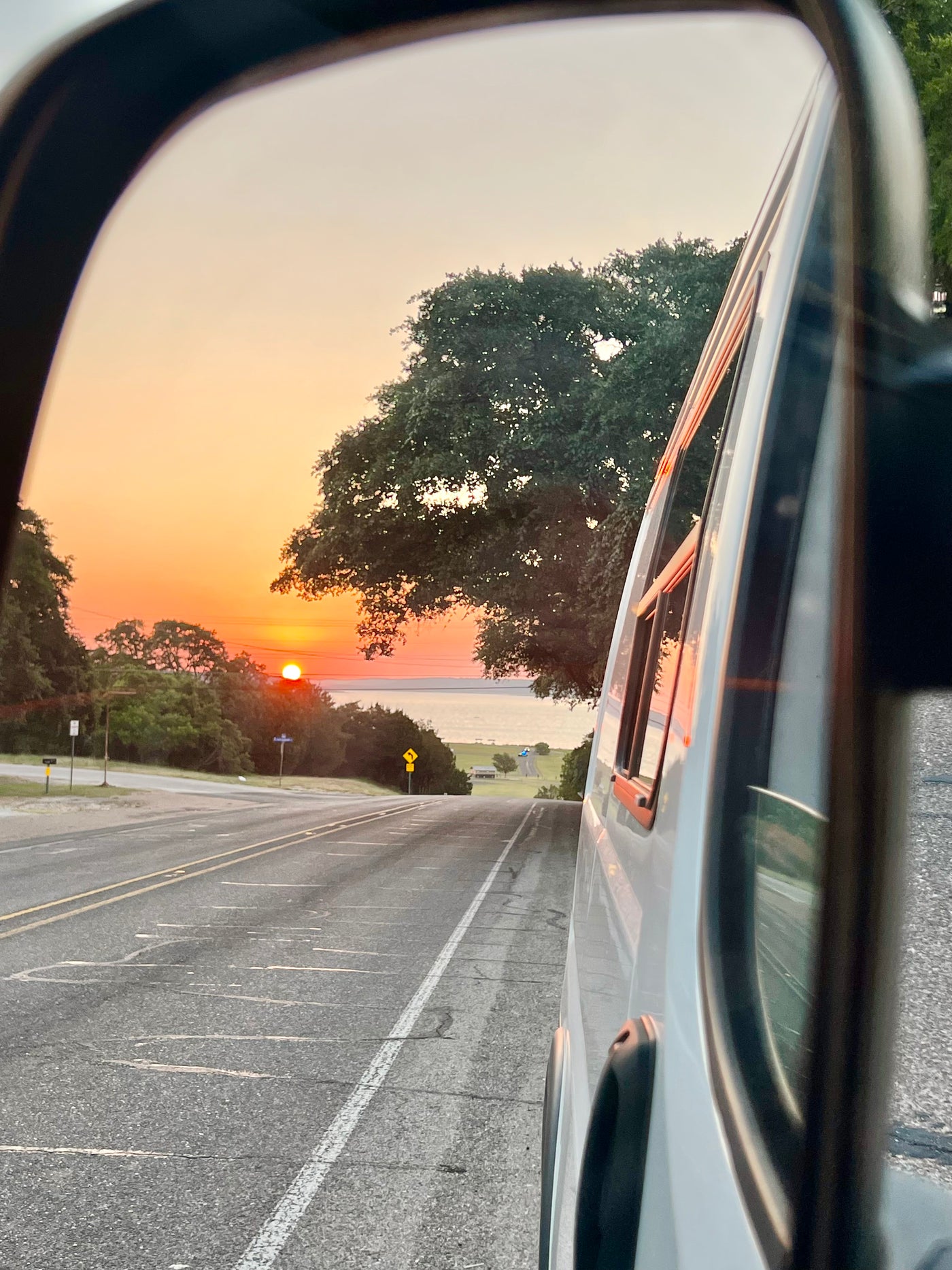 RV rental side mirror reflects a fiery sunset over a lake, with a road and trees. The orange sun hangs low above the horizon. — photo 10