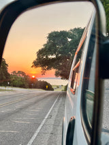 RV rental side mirror reflects a fiery sunset over a lake, with a road and trees. The orange sun hangs low above the horizon.