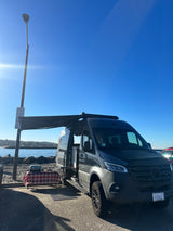 Enjoy an RV rental experience: a dark Sprinter camper van with an awning, table, & grill by the ocean.