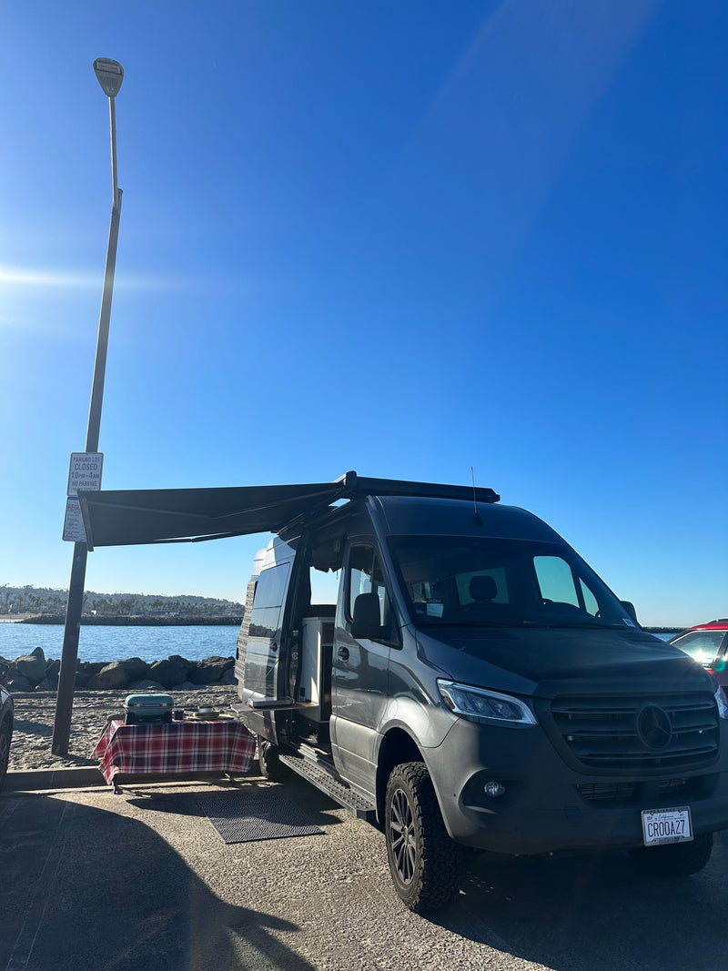 Enjoy an RV rental experience: a dark Sprinter camper van with an awning, table, & grill by the ocean. — photo 11