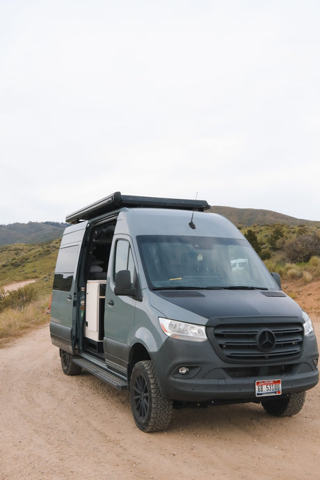 Gray Mercedes Sprinter camper van rv rental on a dirt road with its side door open, revealing interior features for adventure.