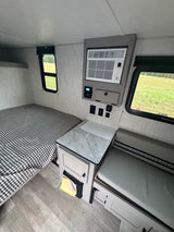 This RV rental's interior boasts a comfortable bed, wall-mounted AC unit, dinette, and bright windows with green outdoor scenery.