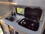 Compact RV rental kitchen with sink, faucet, 2-burner stove (cover open), and a window view of green grass. Ready for meals on the go!