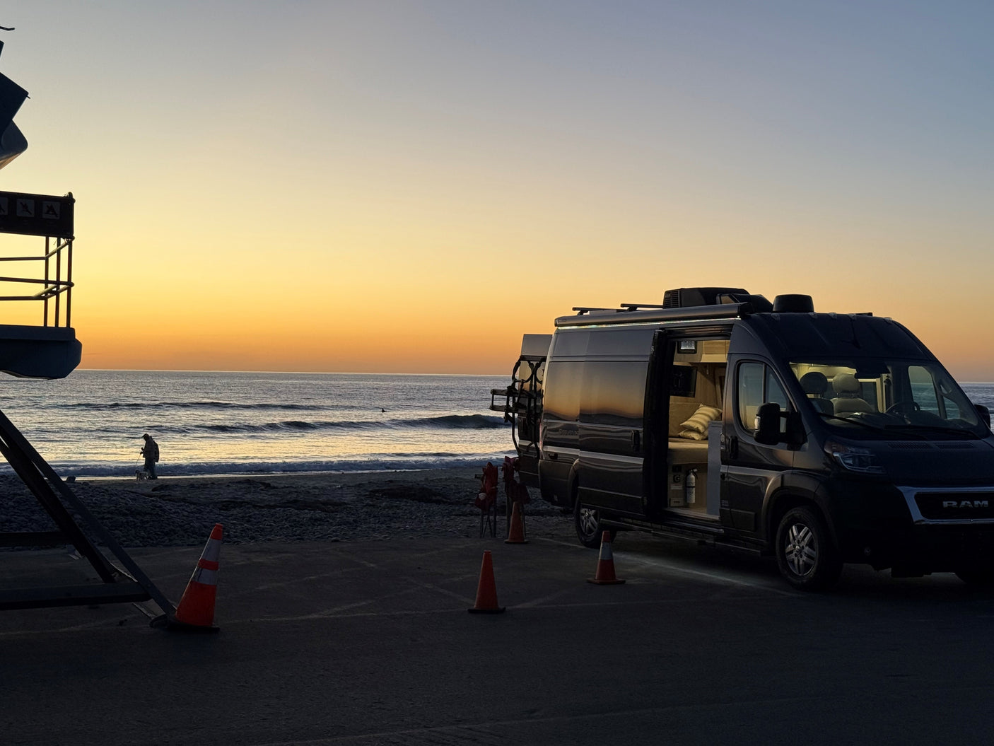 RV rental parked beachside at sunset. Dark camper van with open door, cozy interior. Ocean, lifeguard stand, and person with dog.