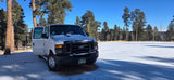 A white Ford van, an RV rental option, parked in snow among pine trees under a clear blue sky.