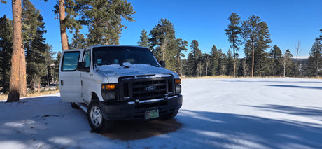 A white Ford van, an RV rental option, parked in snow among pine trees under a clear blue sky.