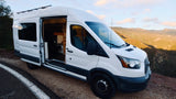 White camper van with open door, showing custom interior, parked on a scenic mountain road at sunset. Ideal for an **rv rental** adventure!