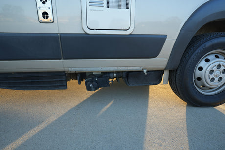RV rental: Side view showing waste tank plumbing, tire, and appliance vents. Sunlit paved surface below.