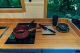 An RV rental's compact kitchen. Bamboo counter, induction cooktop, pot, knife, and dishes by a window overlooking trees.
