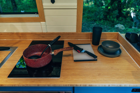 An RV rental's compact kitchen. Bamboo counter, induction cooktop, pot, knife, and dishes by a window overlooking trees.