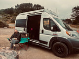 Man relaxes in a chair beside a silver Ram ProMaster RV rental on a gravel road, surrounded by trees.