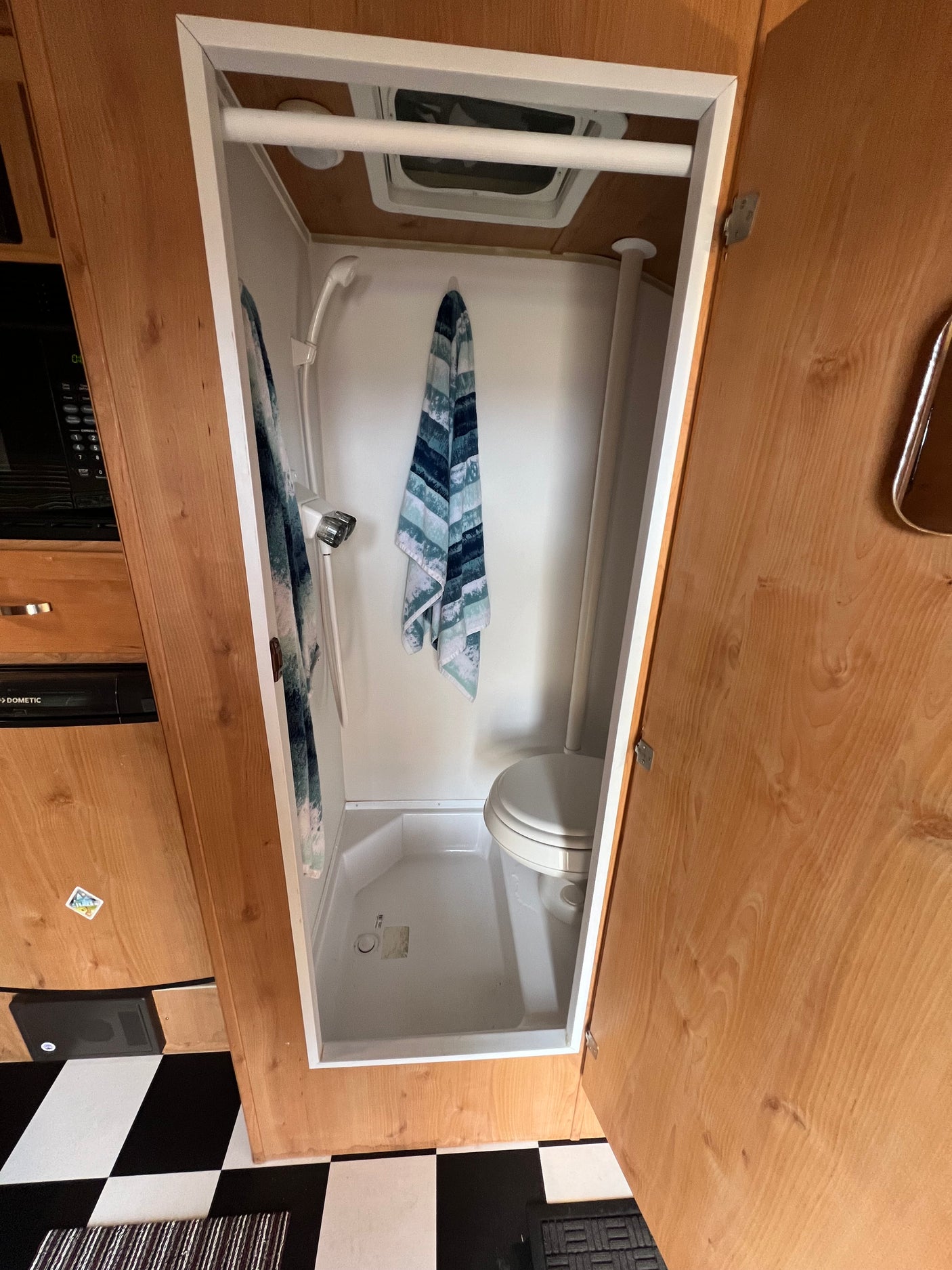 View inside a compact RV rental bathroom, featuring a shower, toilet, ceiling vent, and checkered floor.