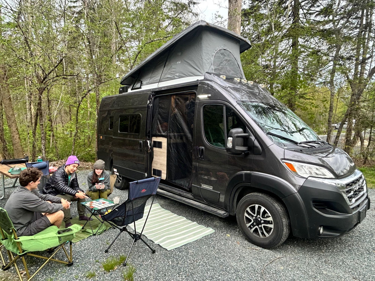 Three people play a board game outside their pop-top RV rental camper van at a wooded campsite.|This RV rental offers a spacious bed and an immersive outdoor view from the open rear doors