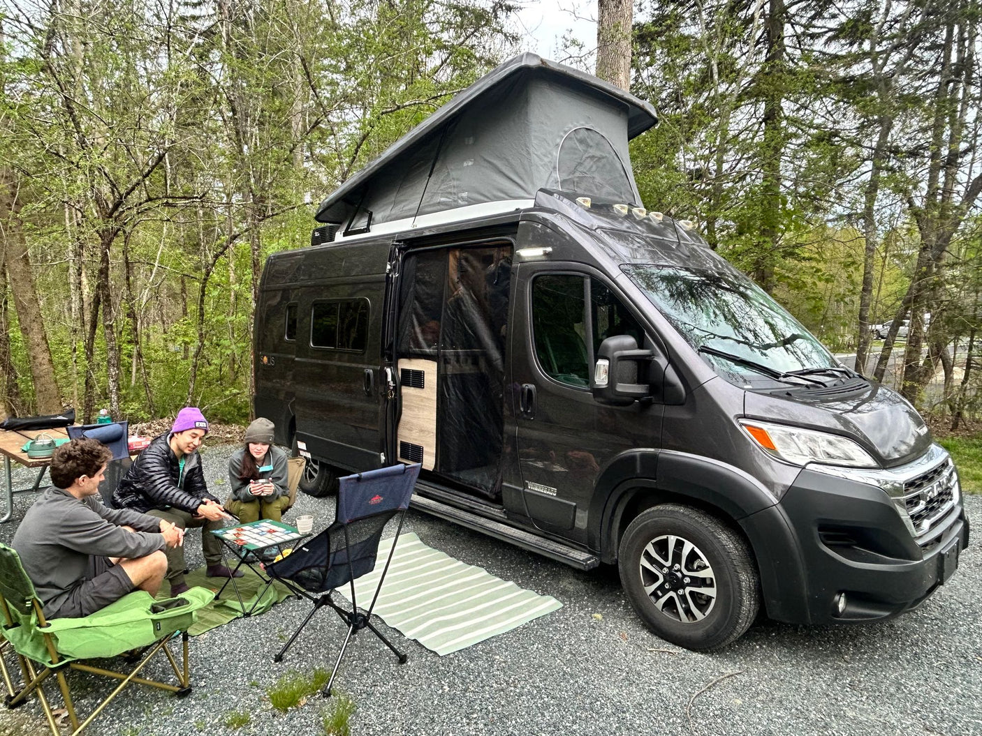 Three people play a board game outside their pop-top RV rental camper van at a wooded campsite.