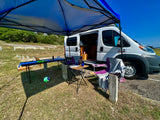 White camper van RV rental set up under a blue canopy with outdoor tables, chairs, and a volleyball, enjoying a sunny day outdoors.