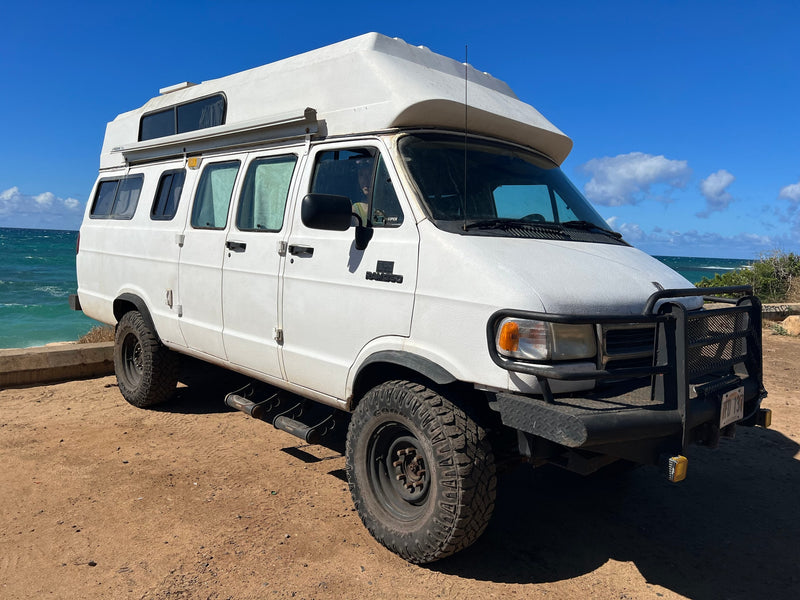  parked on a dirt lot by the blue ocean.|White RV rental adventure van with off-road tires and a raised roof — photo 4