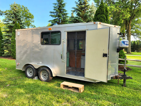 Unique RV rental: a converted silver cargo trailer camper with an open side door & propane tank, parked on grass.