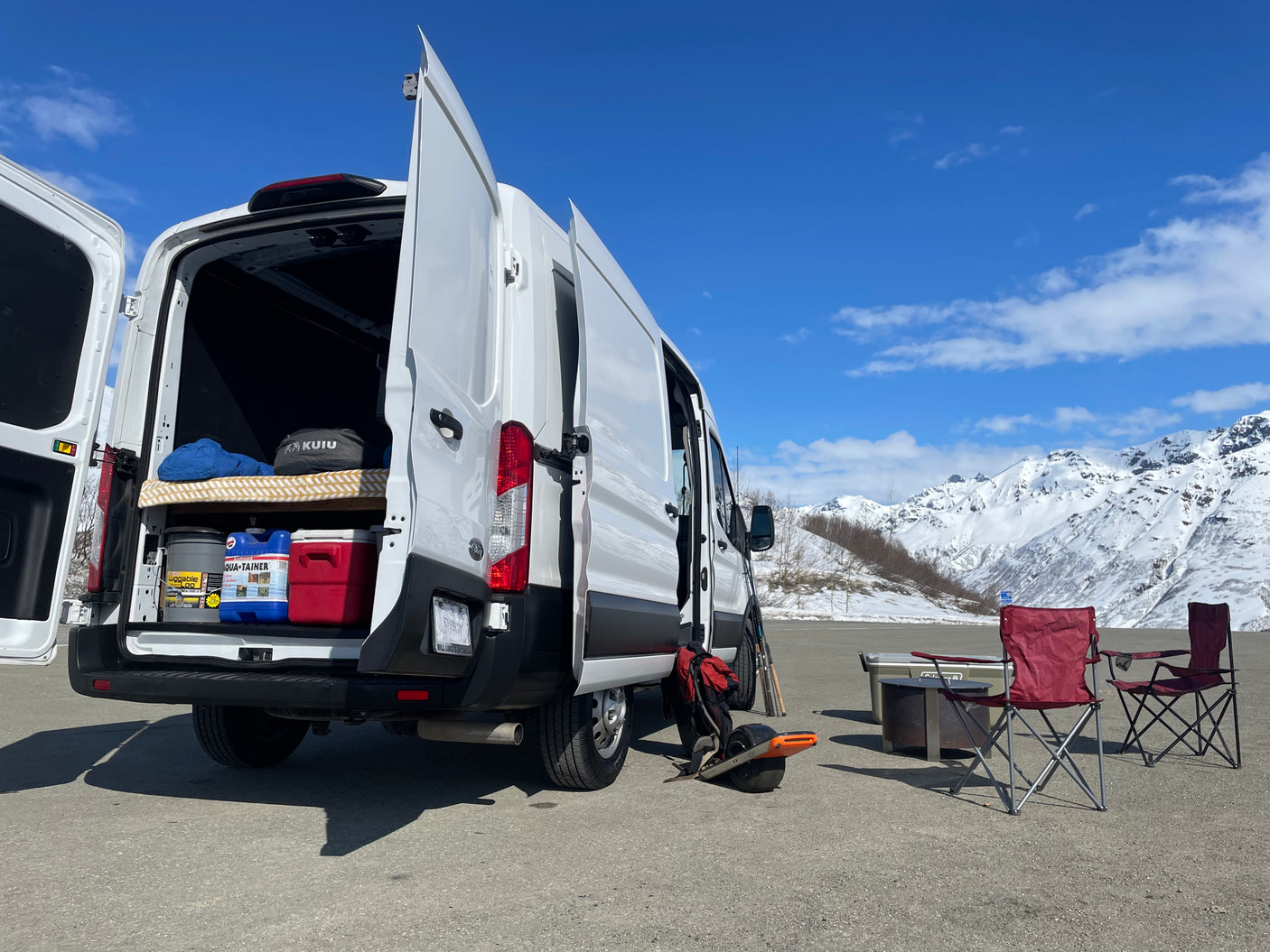 White RV rental van open for a snowy mountain camp, featuring cool gear: cooler, chairs, & Onewheel for adventure.